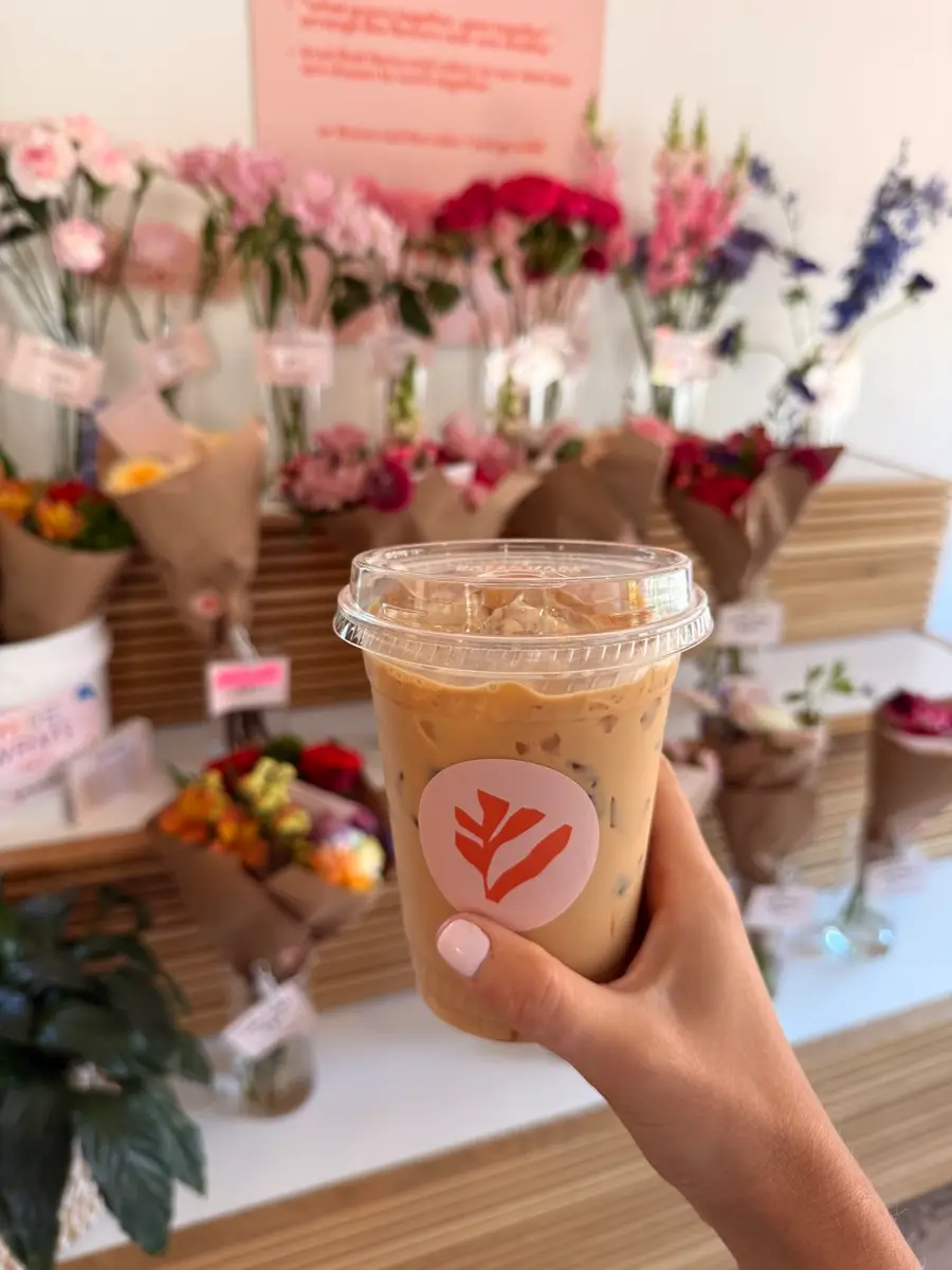 An iced latte held up in front of rows of flowers at the flower market