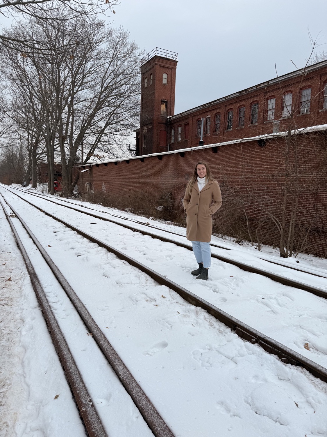 Sally in a camel coat on the snowy rail line by the old mill